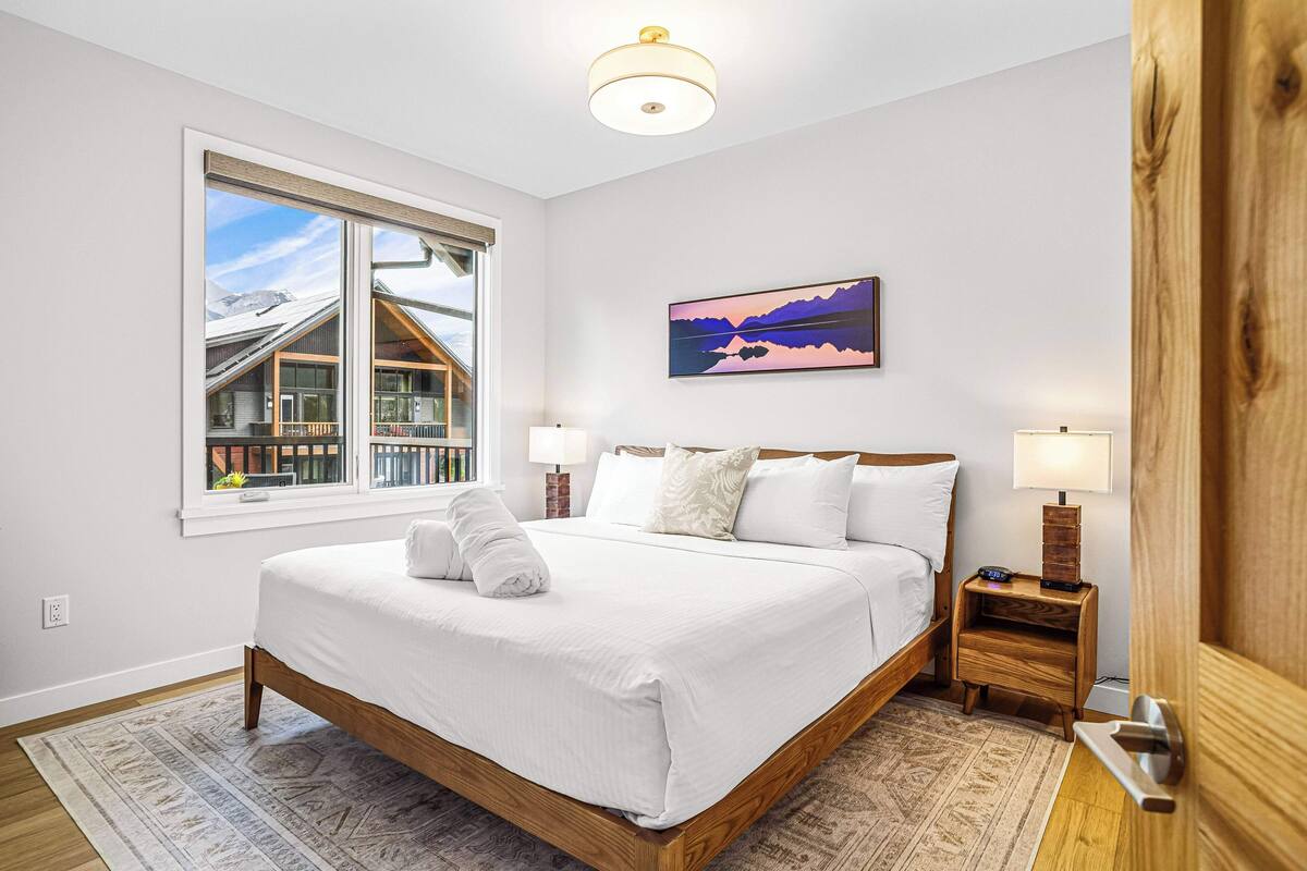 A bedroom with a large bed, a window with a view of a building, a painting on the wall, and a nightstand with a lamp.