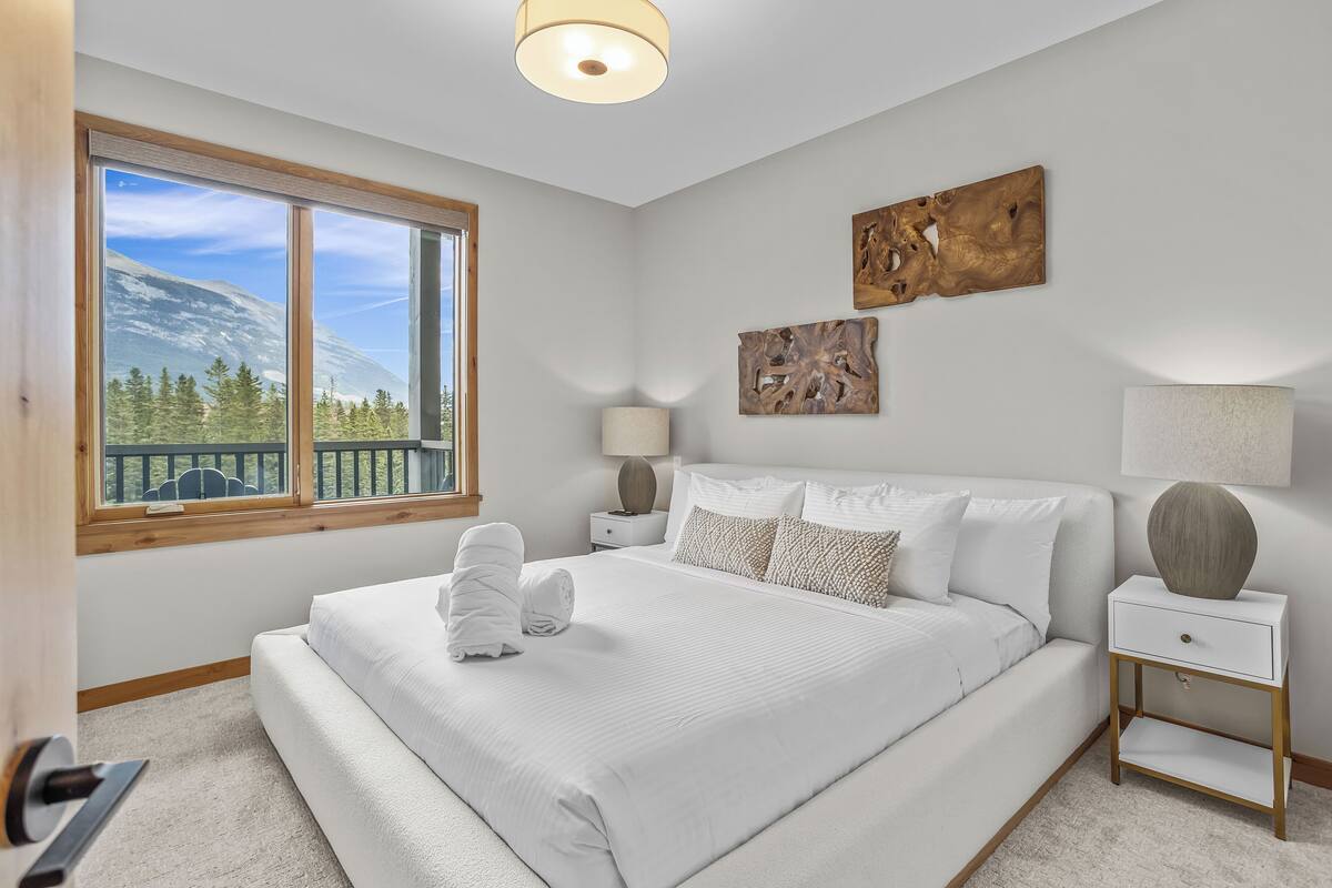 A bedroom with a large bed, a nightstand, a lamp, and a window with a mountain view.