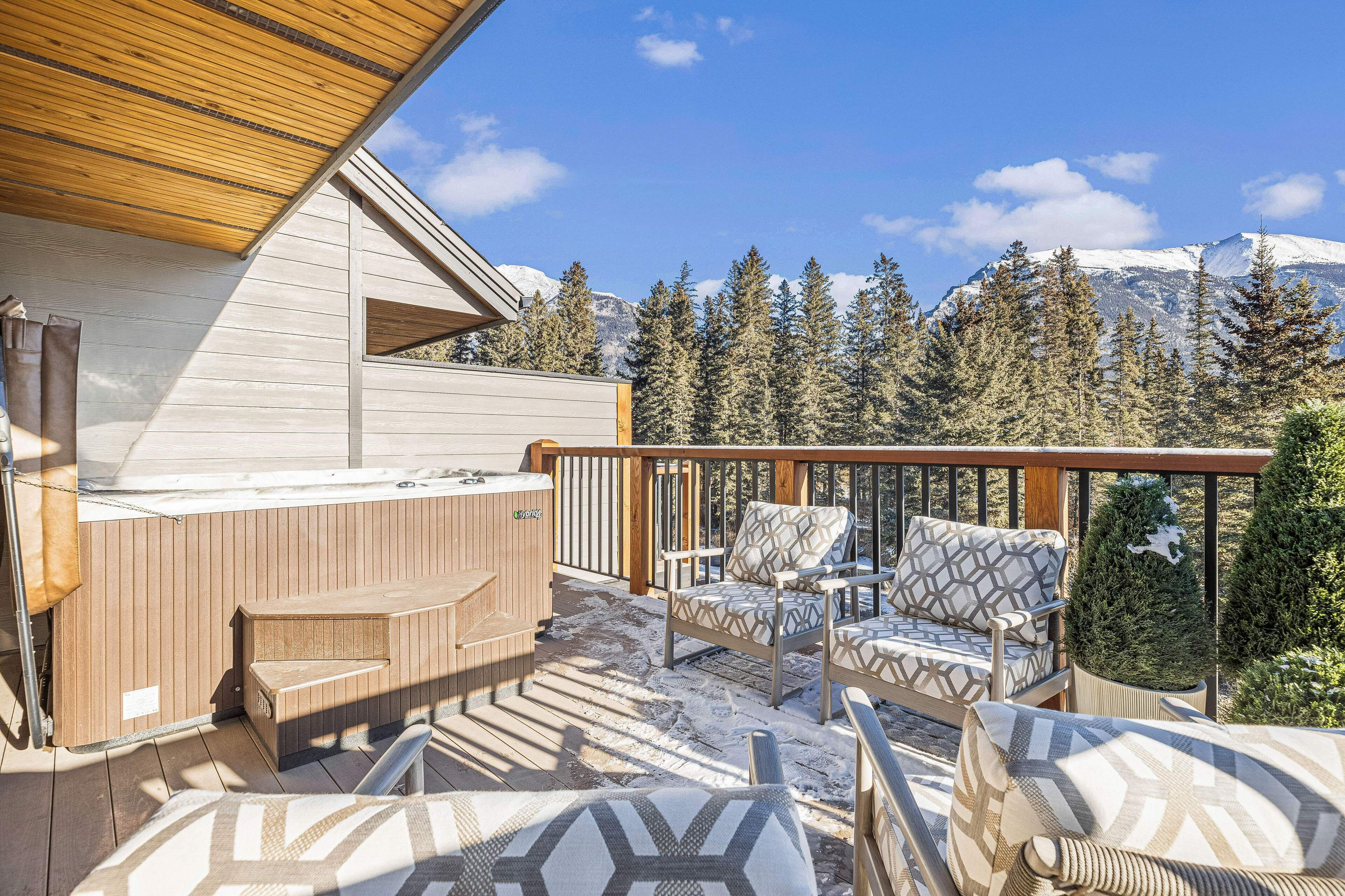A wooden deck with outdoor seating and a hot tub, overlooking a snowy mountain and evergreen trees.