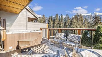 A wooden deck with outdoor seating and a hot tub, overlooking a snowy mountain and evergreen trees.