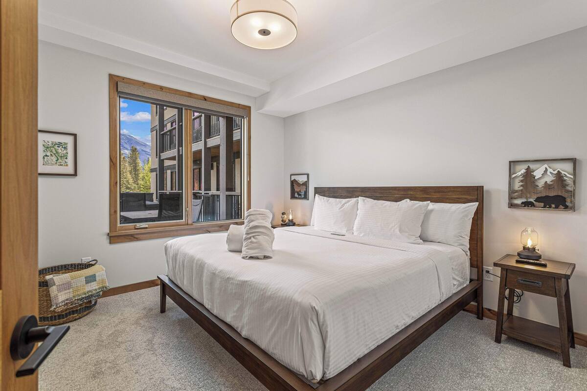 A bedroom with a large bed, a bedside table with a lamp, a window with a view of a building, and a framed picture on the wall.