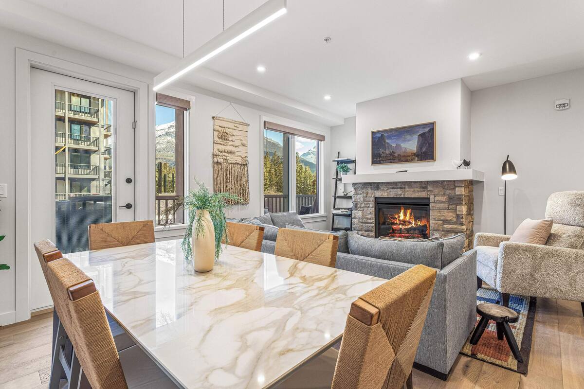 A modern living room with a fireplace, a dining area with a marble table, and a view of mountains through large windows.