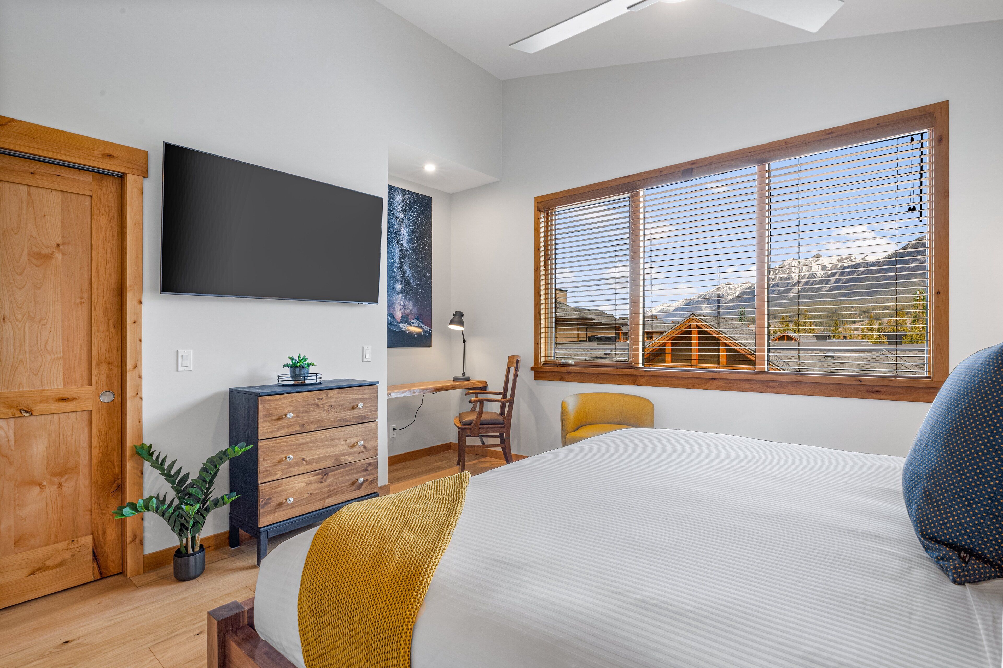 Quarto com cama, escrivaninha, cadeira, televisão, janela com vista para as montanhas e porta de madeira.