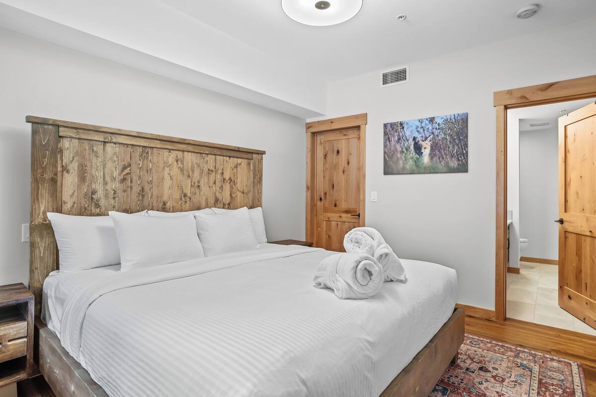 A bedroom with a wooden headboard, white bedding, a rolled towel, and a patterned rug.