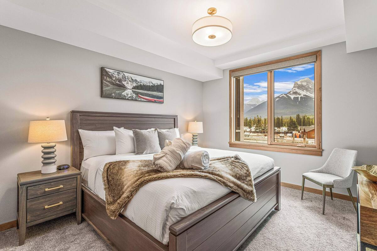 A bedroom with a large bed, two bedside tables with lamps, a chair, a window with a mountain view, and a painting on the wall.