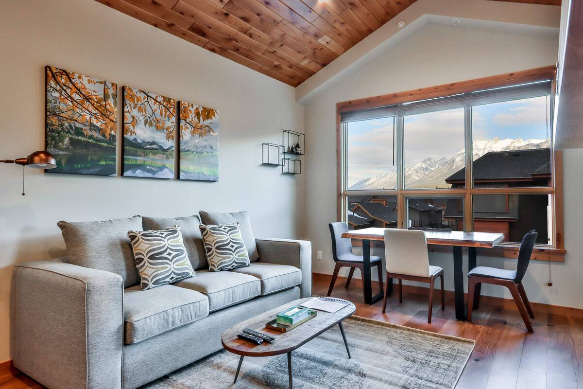 Una sala de estar moderna con un sofá gris, una mesa de centro y una zona de comedor con una mesa y sillas. Hay tres cuadros enmarcados en la pared y un ventanal con vistas a la montaña.
