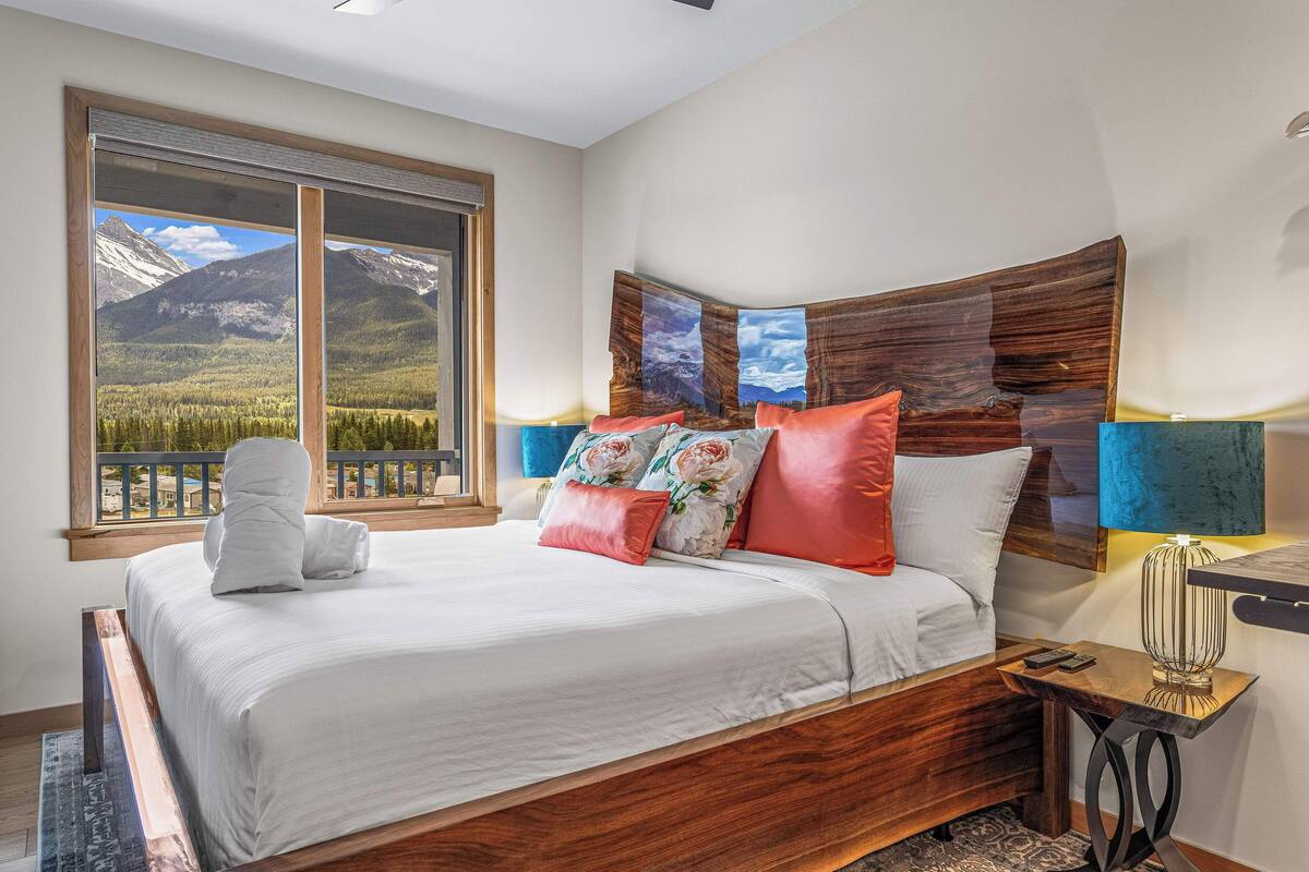 A bedroom with a large window offering a mountain view, a wooden headboard, and a bedside table with a lamp.