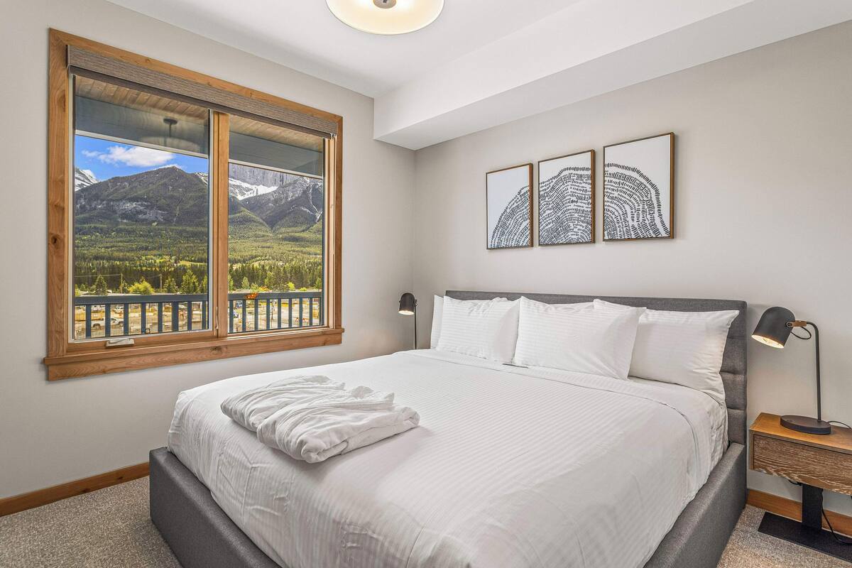 A bedroom with a large bed, a view of mountains through a window, and two framed artworks on the wall.