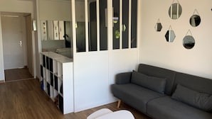 Moderni olohuone, jossa on harmaa sohva, valkoinen pöytä ja valkoinen kaappi lasiovilla. Seinällä on peilejä ja ovi johtaa toiseen huoneeseen.