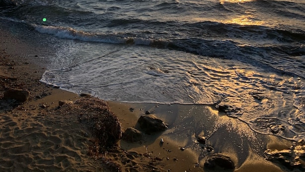 Ranta, jossa aallot lyövät rantaviivaan auringonlaskun aikaan, kivinen rantakaistale ja vasemmalla puolella oleva rakennus.