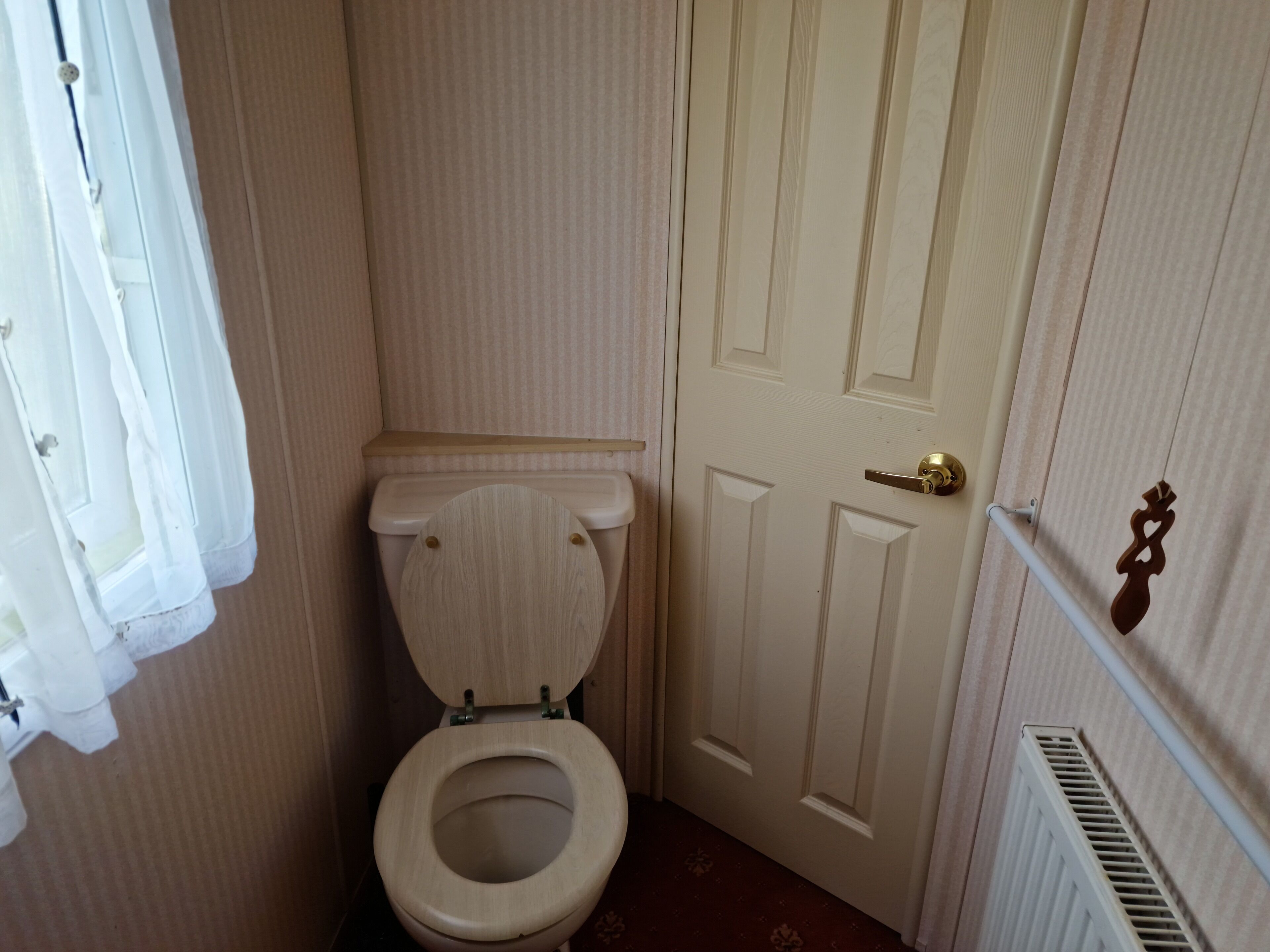 A small, compact bathroom with a wooden toilet, a white door with a brass handle, and a radiator on the right.
