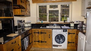A compact kitchen with wooden cabinetry, a built-in washing machine, and a window overlooking a garden. - Secluded, rural retreat nestled in Sherwood Forest, with hot tub (Newark)