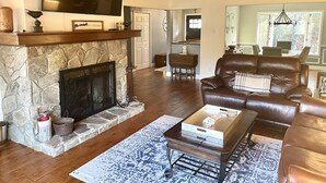 A spacious living room with a stone fireplace, leather sofa, and a flat-screen TV mounted above it. - Direct Lakefront Connestee Falls: Views + private dock + 4 kayaks + Not steep! (Brevard)
