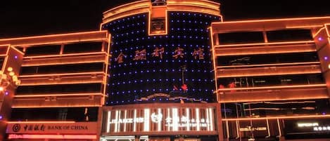 Un edificio de varios pisos con señalización iluminada y una entrada destacada, rodeado de automóviles estacionados.