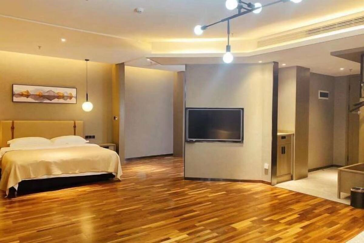 Una habitación de hotel moderna con suelo de madera, una cama con ropa de cama blanca, un televisor de pantalla plana montado en la pared y una lámpara en el techo.