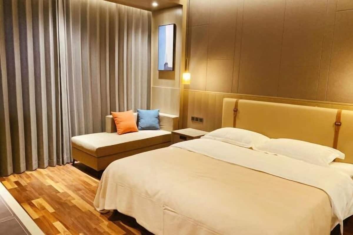 Habitación de hotel con una cama grande, una zona de descanso más pequeña con cojines, suelo de madera y un amplio ventanal con cortinas.