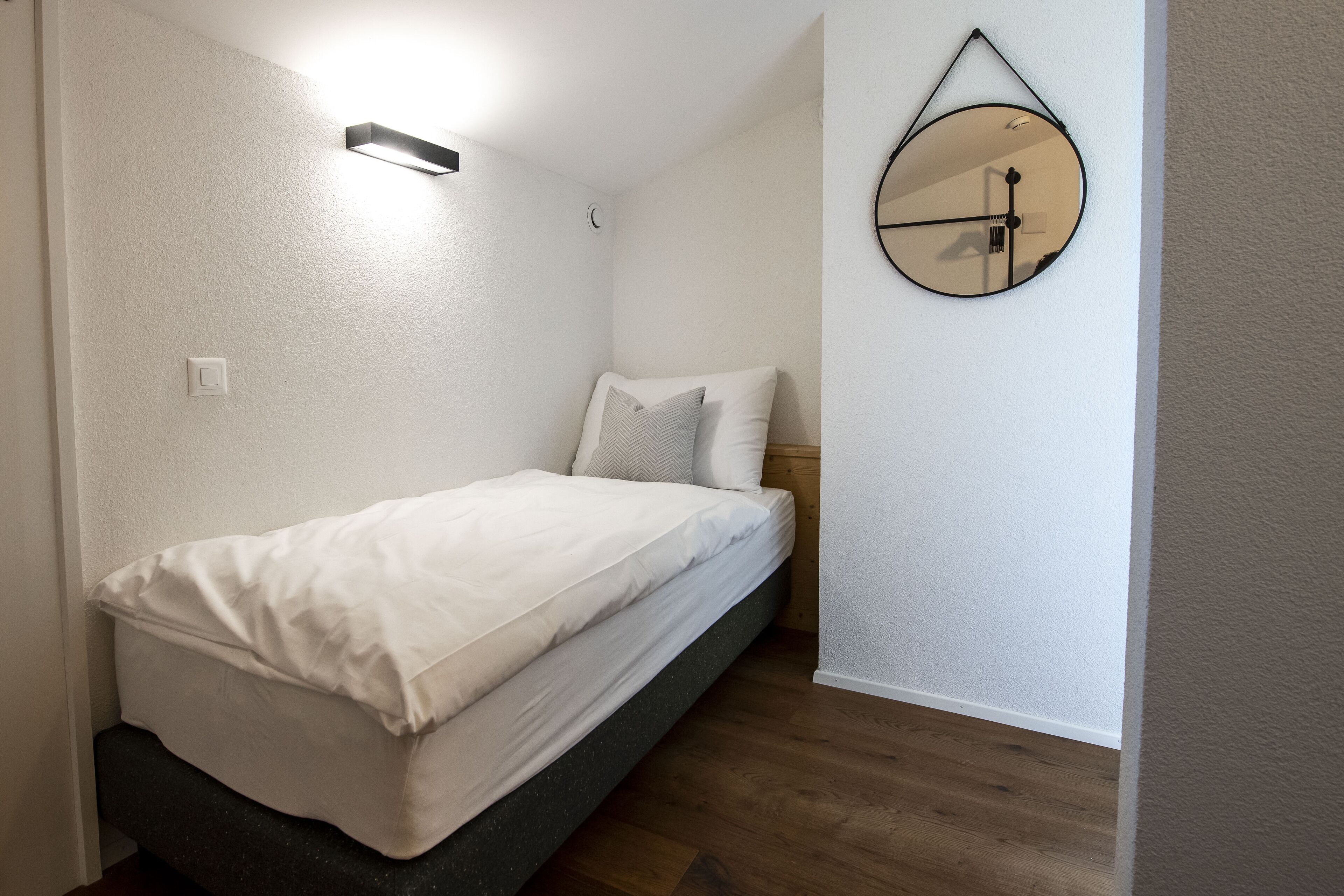 A small, neatly arranged bedroom with a single bed, a wall-mounted mirror, and a light fixture.