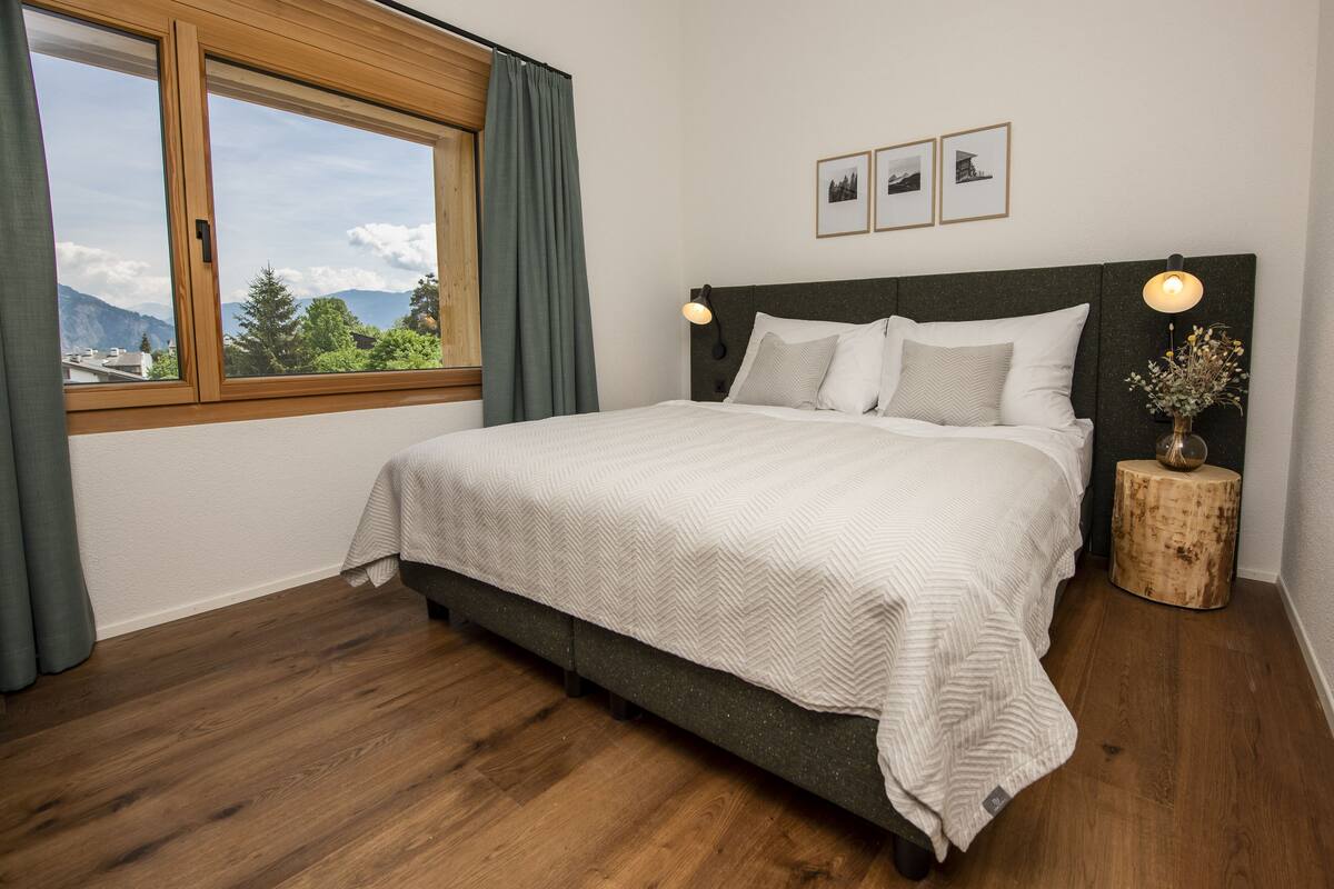 A bedroom with a large bed, wooden headboard, a window with curtains, a bedside table with a lamp, and framed pictures on the wall.