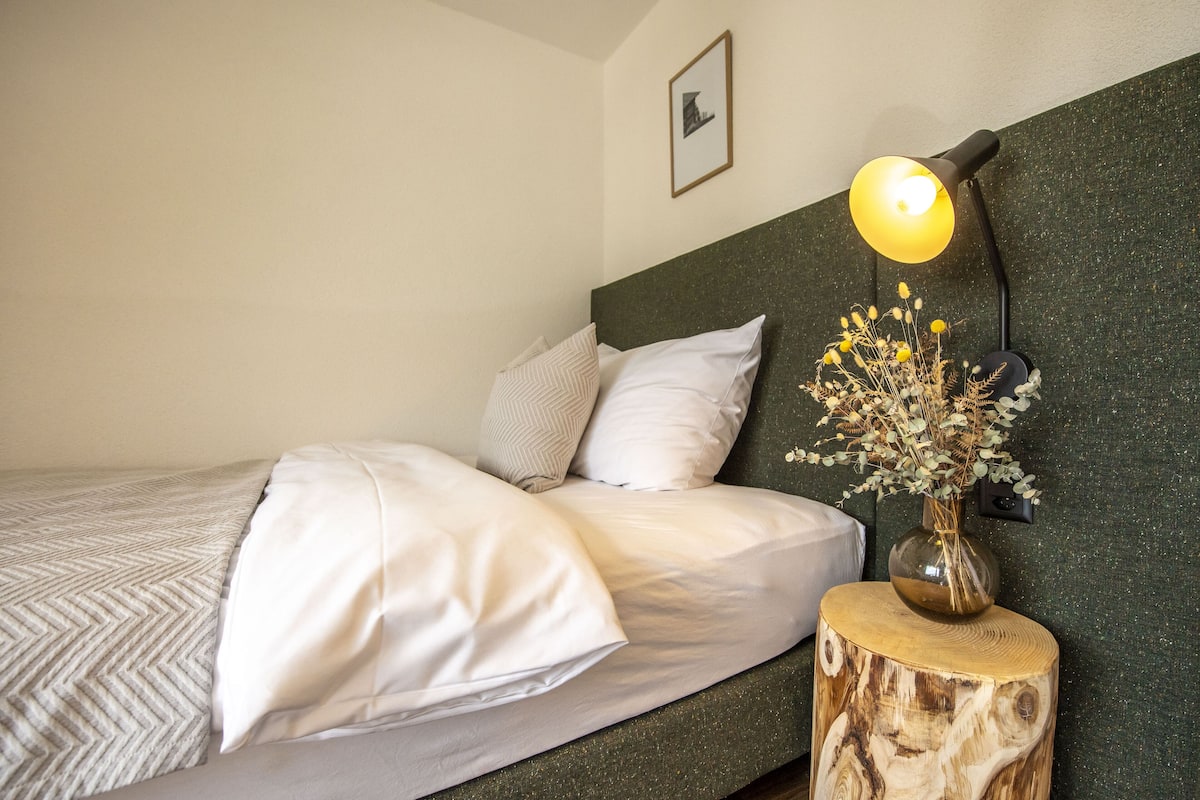 A neatly made bed with white linens and a wooden headboard. A small wooden nightstand with a vase of dried flowers and a lamp is beside the bed.