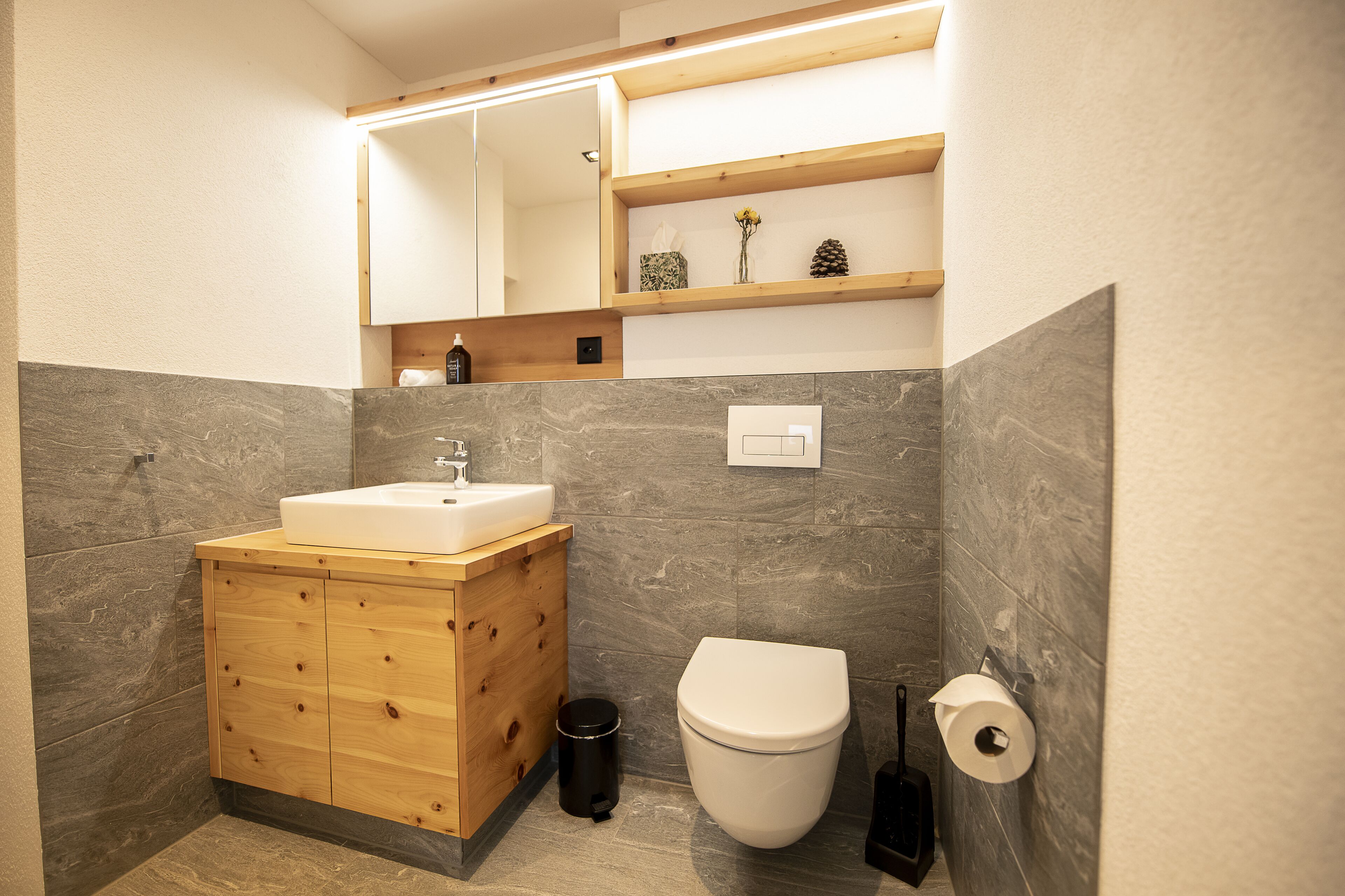 Un baño moderno con tocador de madera, un lavabo blanco, un inodoro y un estante sobre el tocador.