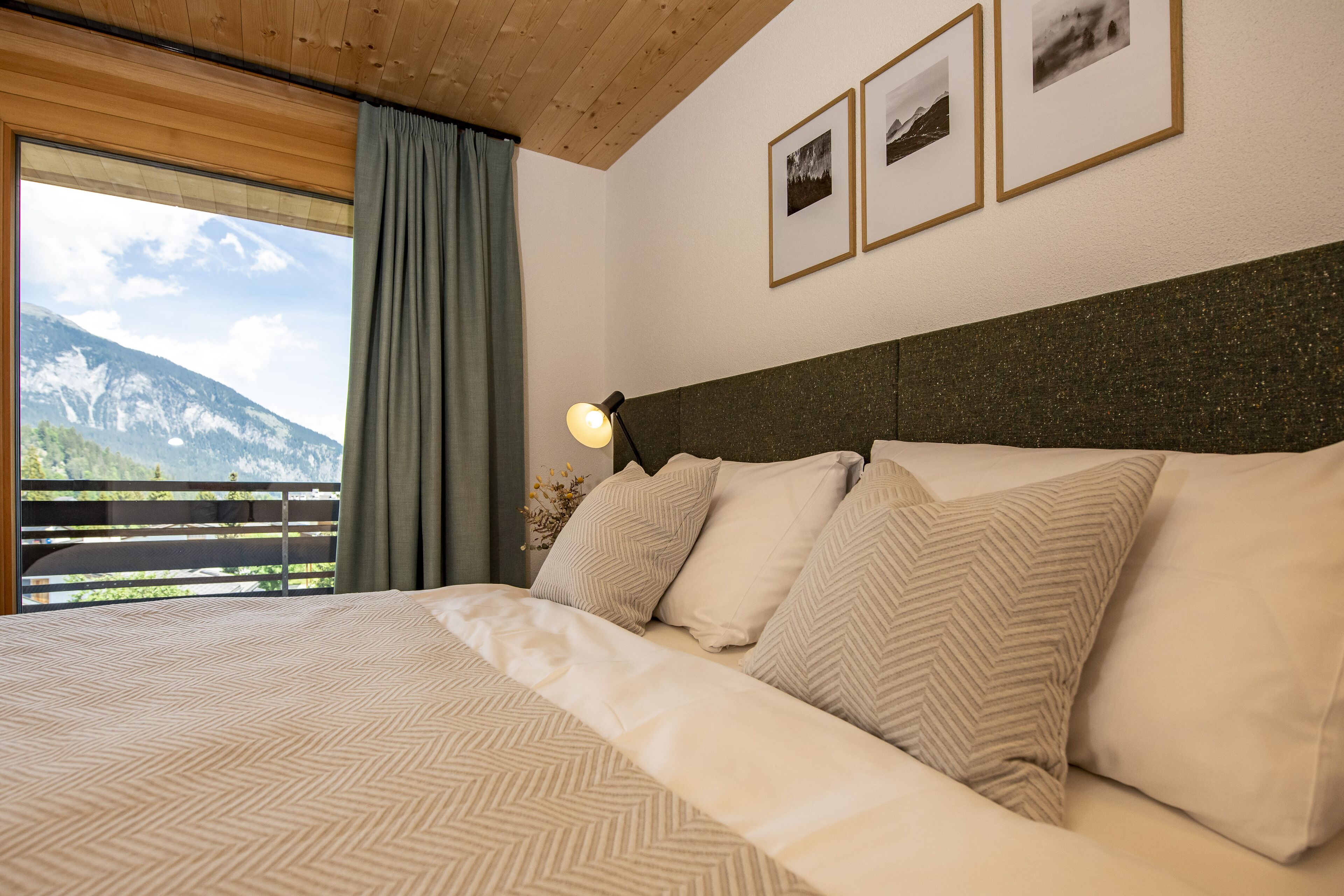 Een slaapkamer met een bed, hoofdeinde, raam met uitzicht op bergen en een lamp.