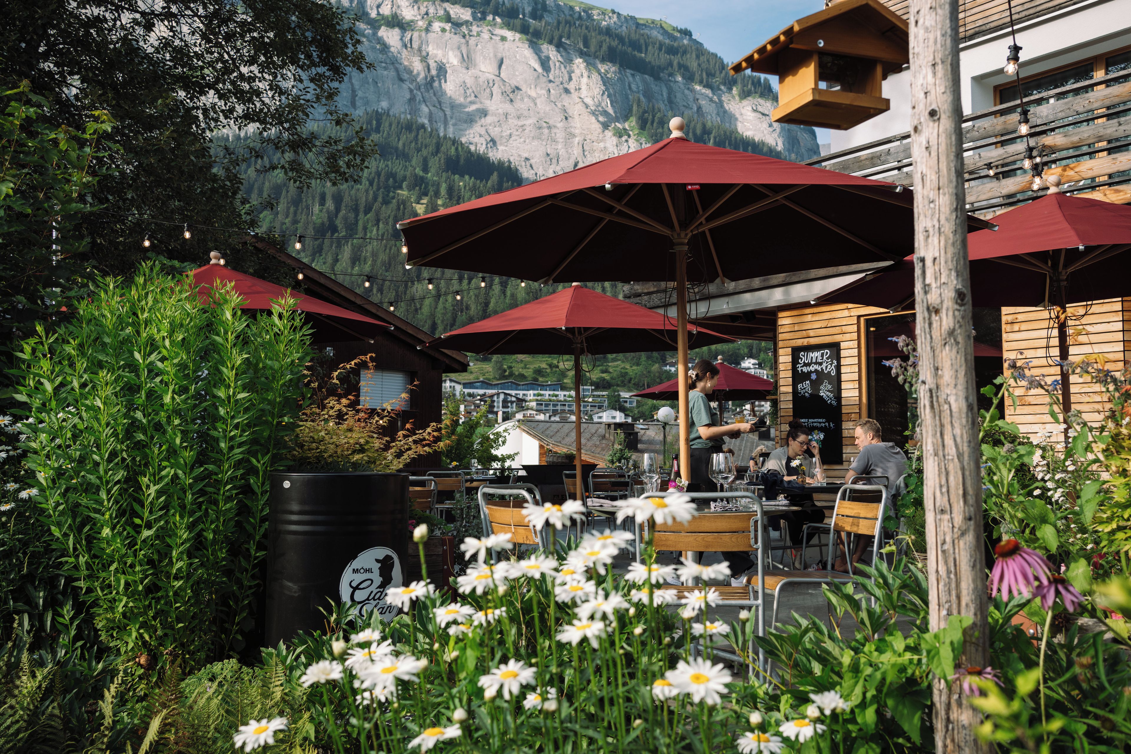 An outdoor café with red umbrellas, wooden tables, and chairs, surrounded by greenery and flowers.