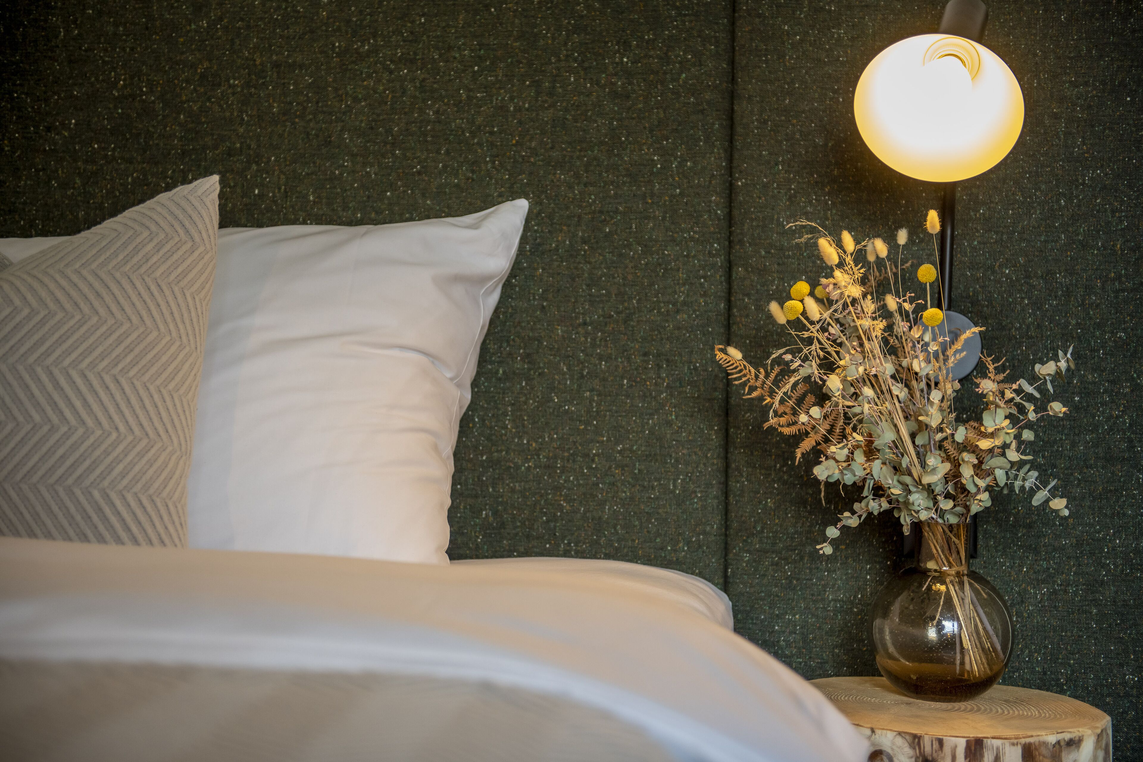 Una cama bien tendida con sábanas blancas y un cojín decorativo, un jarrón con flores secas sobre una mesita de noche de madera, y una lámpara de pared con luz cálida.