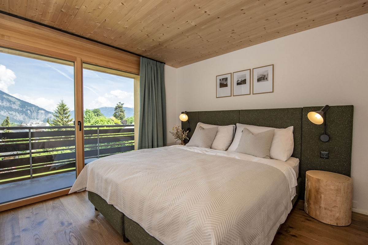 A bedroom with a large bed, wooden ceiling, and a view of mountains through a sliding door.