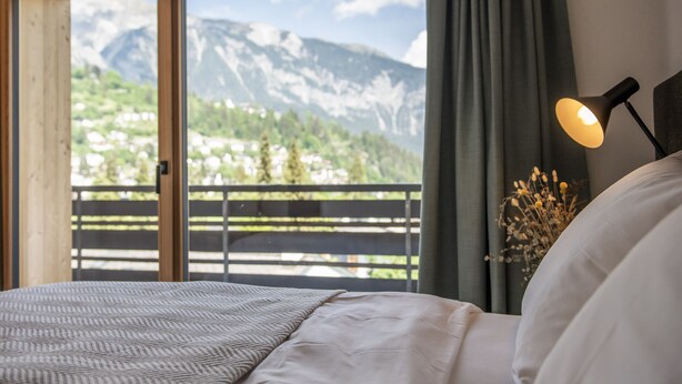 A bedroom with a bed, a nightstand with a lamp, a window with a view of mountains, and a balcony with a railing.
