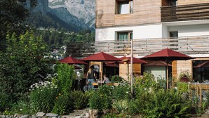 A modern building with a wooden facade, outdoor seating with red umbrellas, and a mountainous backdrop.
