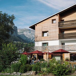 A modern building with a wooden facade, outdoor seating with red umbrellas, and a mountainous backdrop. - Flem Mountain Lodge (Flims)