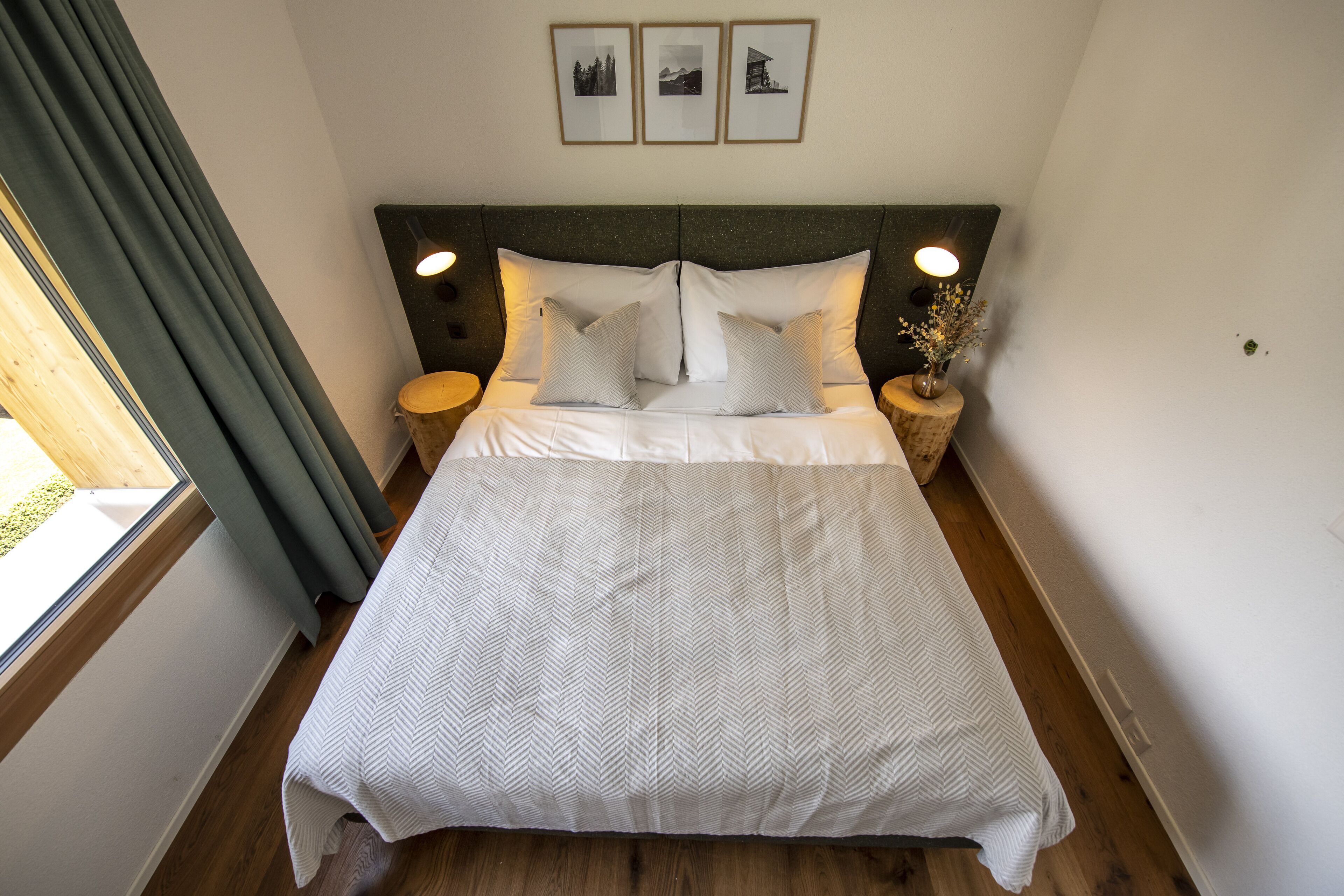 Una cama bien tendida con colcha de estampado romboidal, dos almohadas y cabecera con acabado oscuro. Hay dos mesitas de noche con lámparas y una ventana con cortinas en el lado izquierdo.