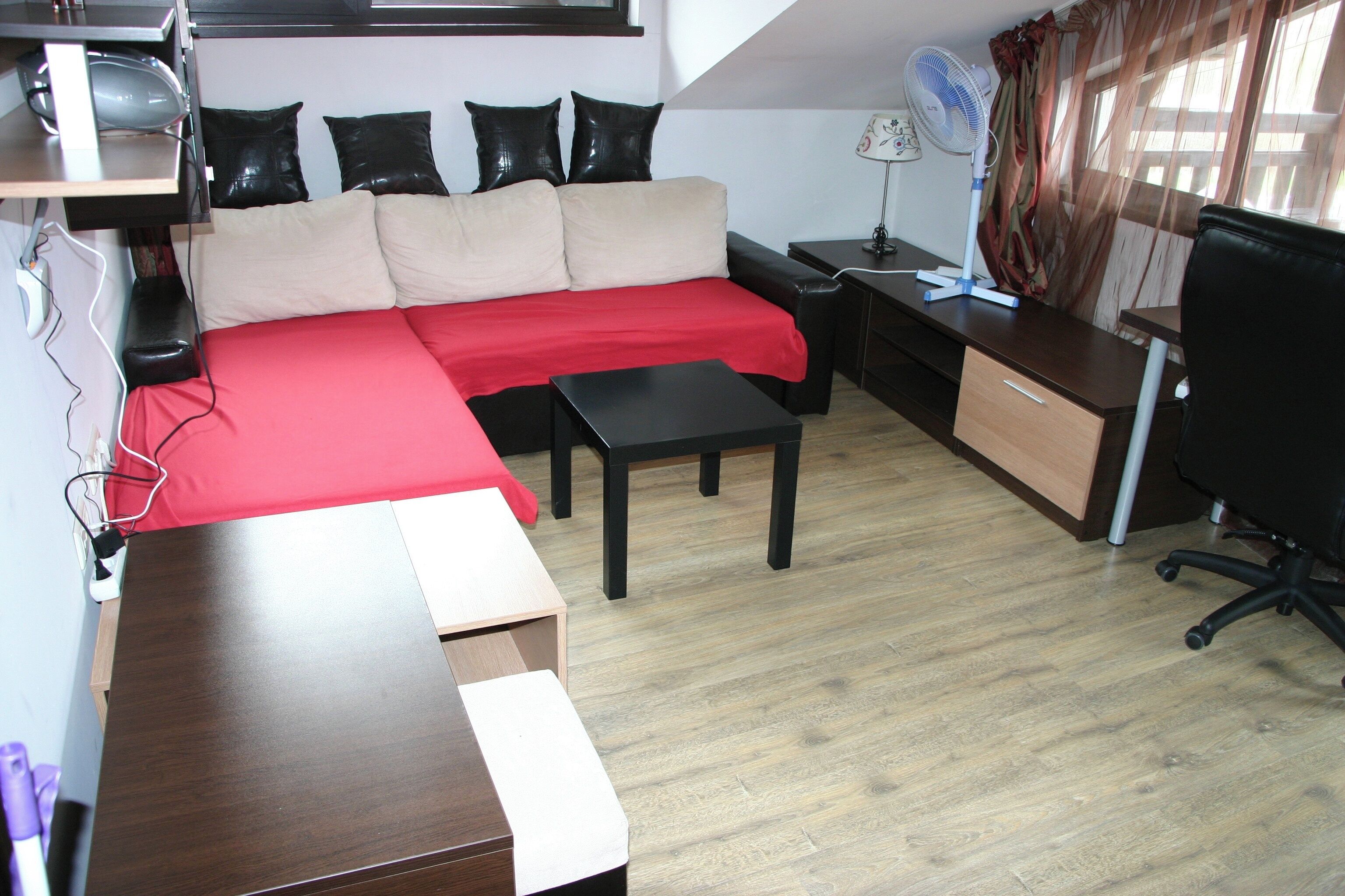 A compact living space with a sofa, a small table, a desk, a chair, a fan, and a TV.