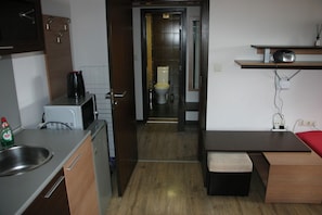 A compact kitchen with a microwave, kettle, and sink; a small dining area with a bench; a toilet in the adjacent room; and a shelf with a radio.