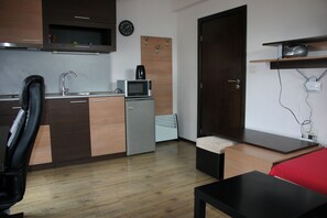 Une cuisine compacte Ă©quipĂ©e dâun four Ă micro-ondes, dâune machine Ă cafĂ© et dâun petit rĂ©frigĂ©rateur. Le salon comprend une porte en bois, une chaise noire et un canapĂ© rouge.