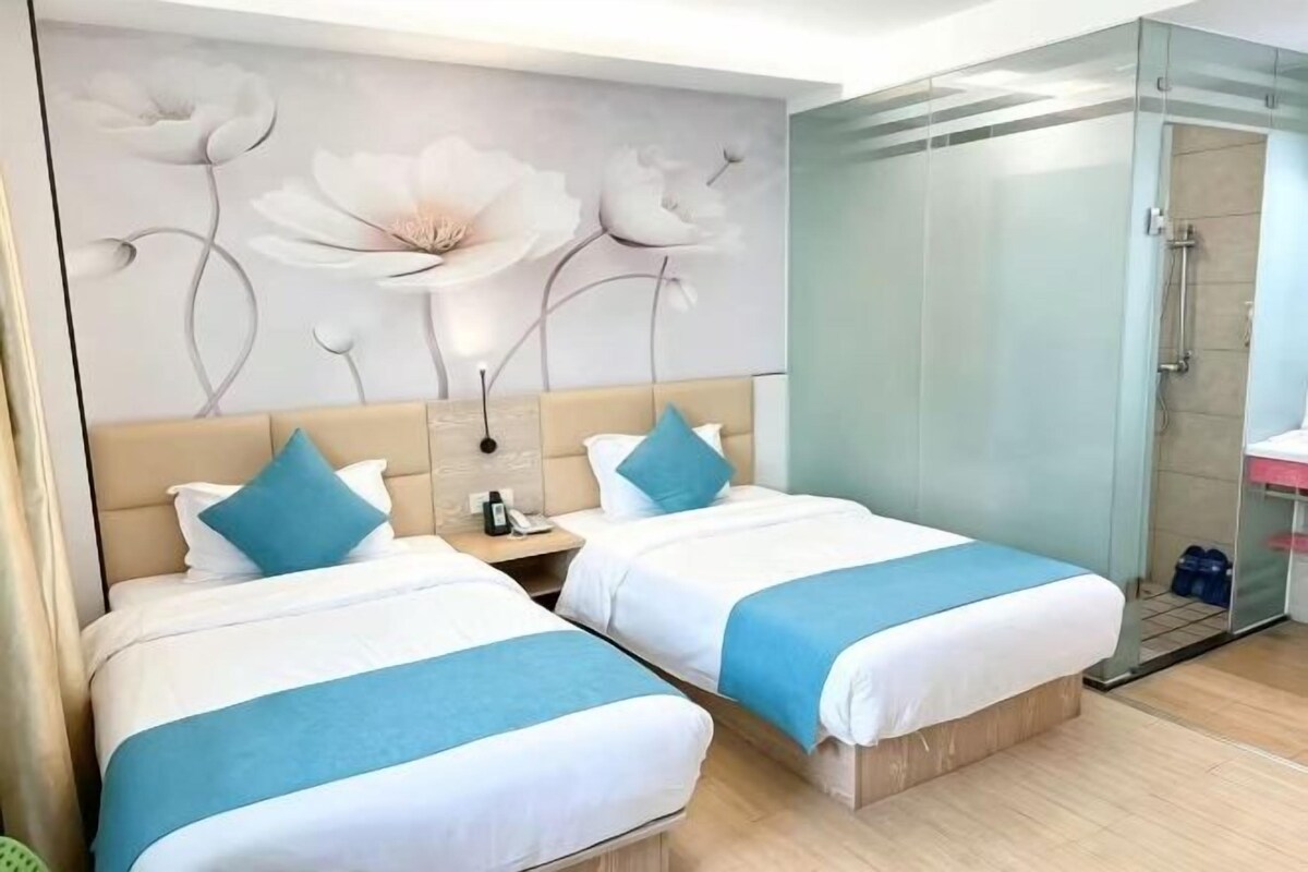 Dos llits amb roba de llit blava i blanca, una paret amb un mural floral, un espai de banyera amb vidre i un bany petit amb un lavabo i un mirall.