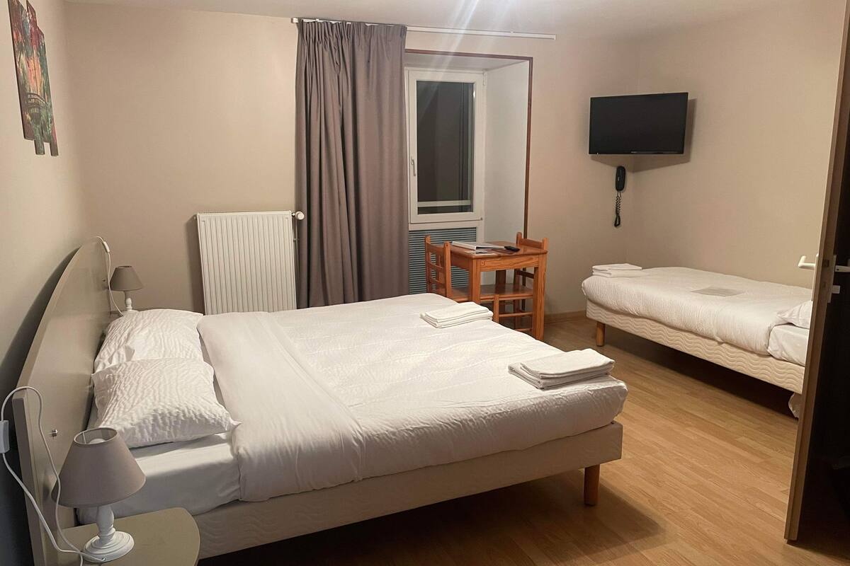A hotel room with a large bed, a smaller bed, a desk with a chair, a TV mounted on the wall, and a window with curtains.