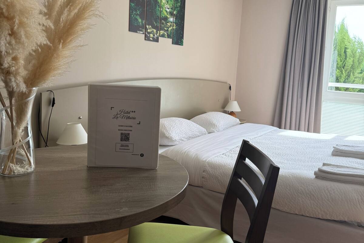 A hotel room with a bed, a desk with a laptop, a chair, a vase with dried plants, and a window with curtains.