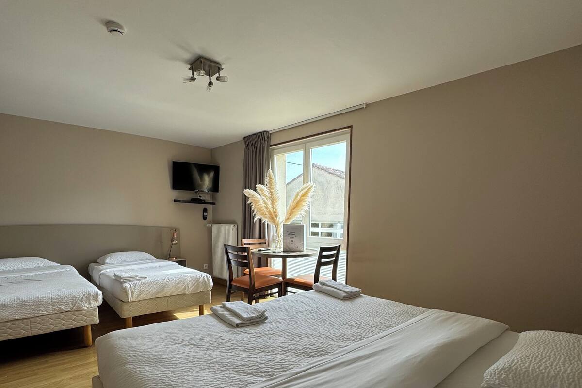 A hotel room with two single beds, a large bed, a dining table with chairs, a TV mounted on the wall, and a window with curtains.