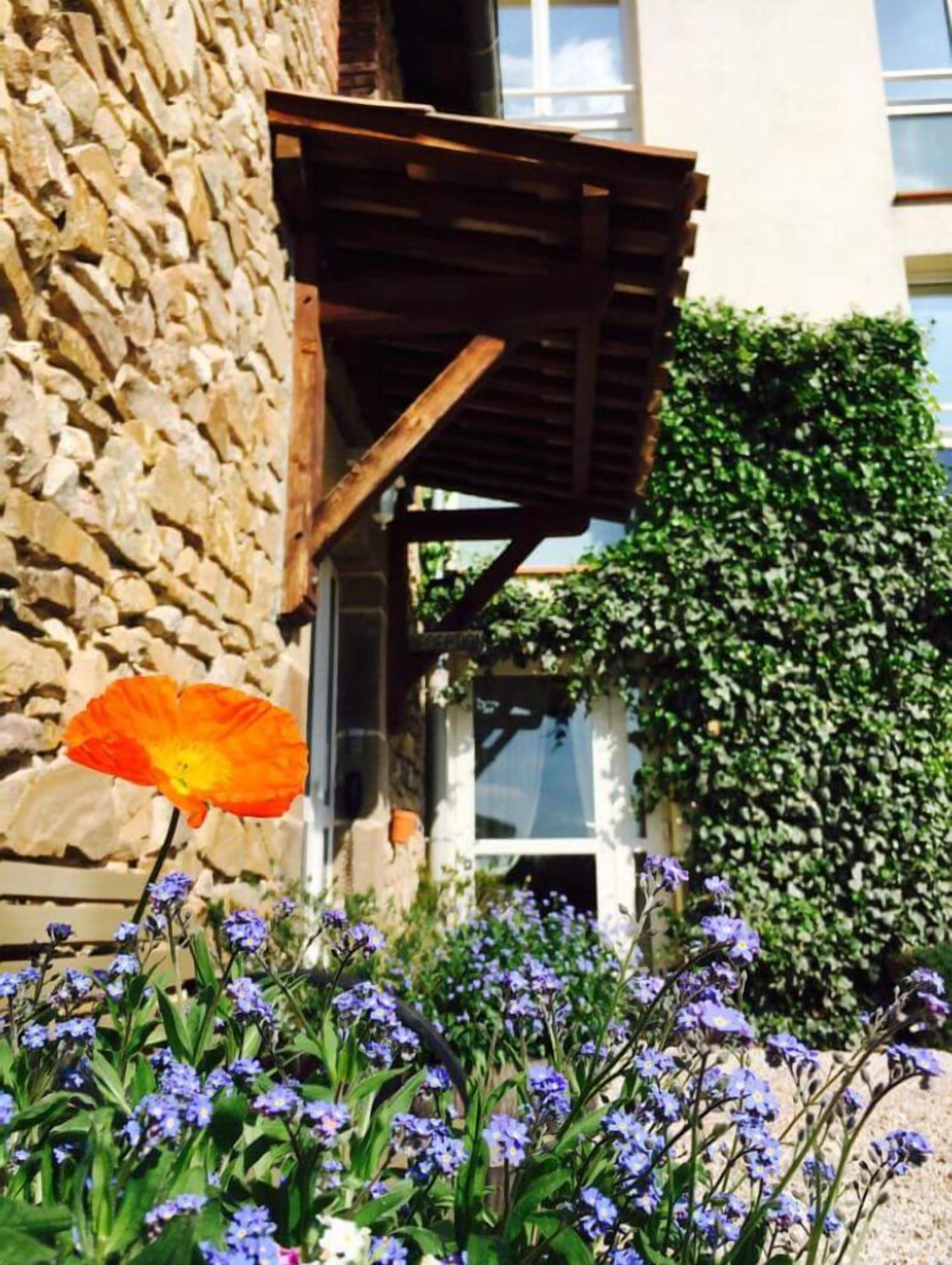 Un edificio de piedra con una pérgola de madera, rodeado de plantas florecientes y un seto verde.