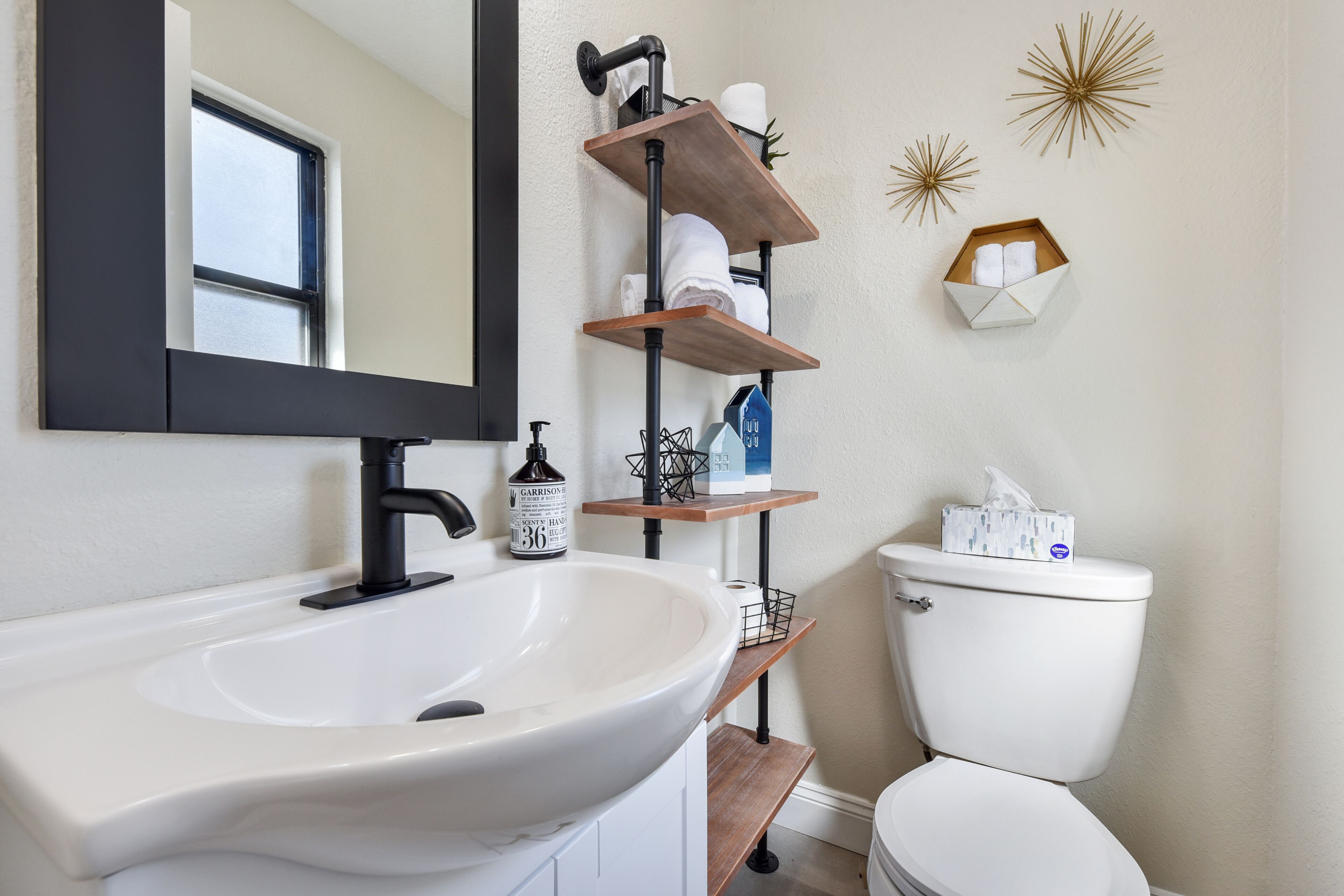 Un baño con un lavamanos blanco, grifo negro y un inodoro. Hay repisas de madera sobre el lavamanos y el inodoro, con toallas y artículos de aseo.