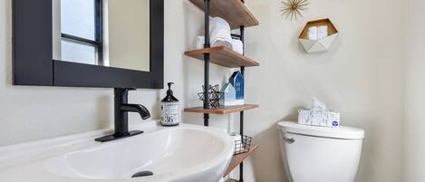 Un baño con un lavamanos blanco, grifo negro y un inodoro. Hay repisas de madera sobre el lavamanos y el inodoro, con toallas y artículos de aseo.