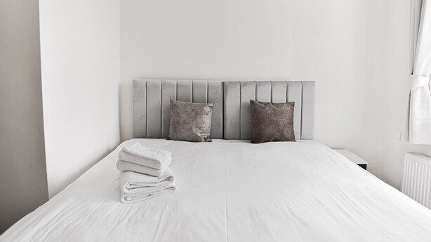 Ein ordentlich bezogenes Bett mit grauem Kopfteil, zwei Kissen und einem Stapel gefalteter Handtücher.