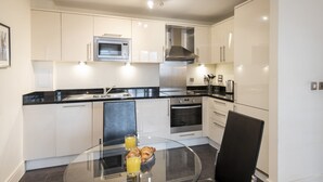 A modern kitchen with white cabinets, a black countertop, a stainless steel microwave, and an oven. There is a glass table with a plate of croissants and a glass of orange juice. - Charming 1-bed Apartment in Great Suffolk Street (London)
