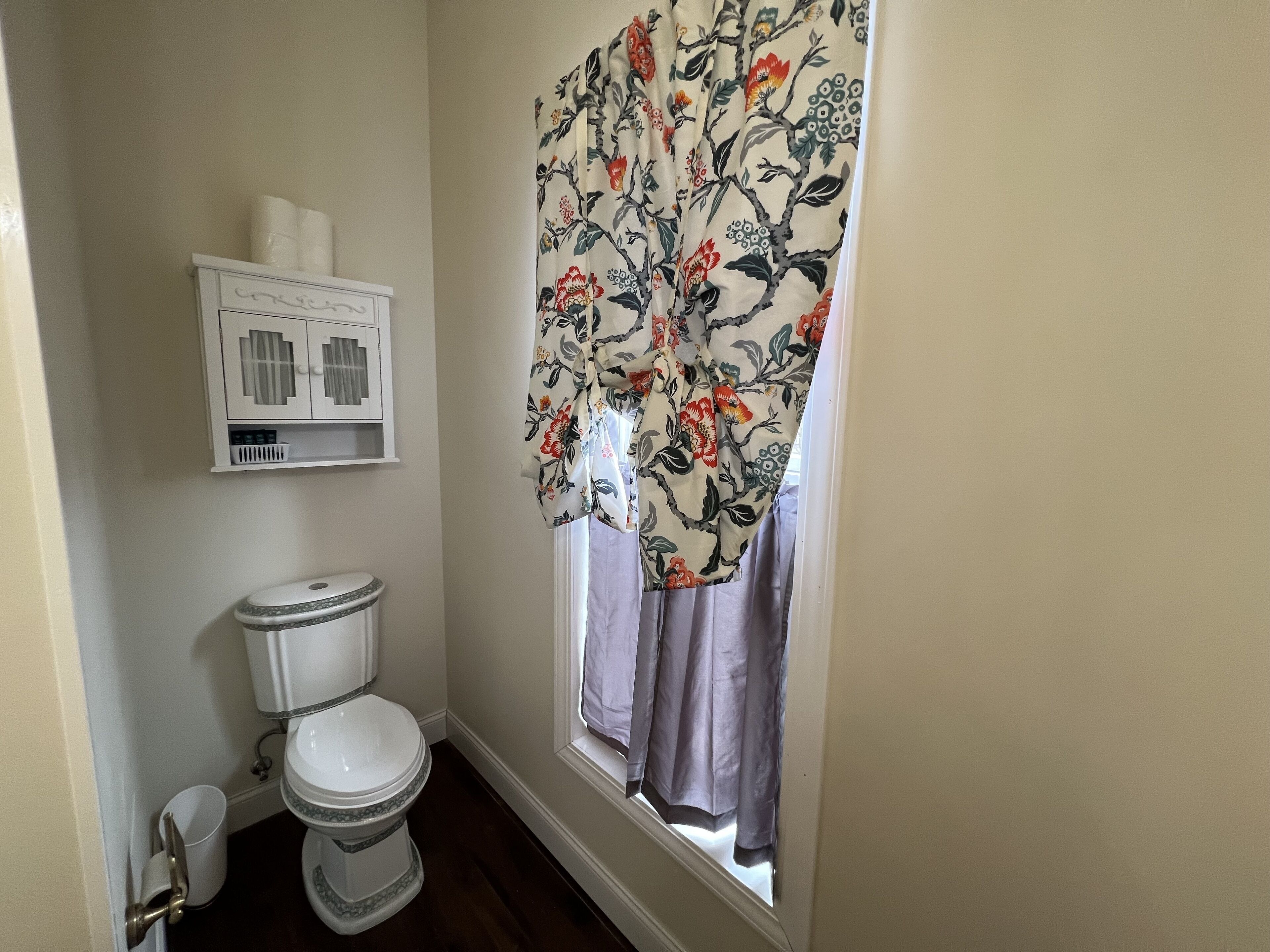Une salle de bain avec des toilettes, un meuble contenant du papier toilette et une fenêtre ornée d’un rideau à motifs floraux.