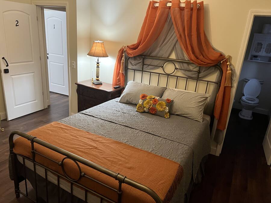 A bedroom with a bed, a nightstand, a lamp, and a bathroom visible through an open door.