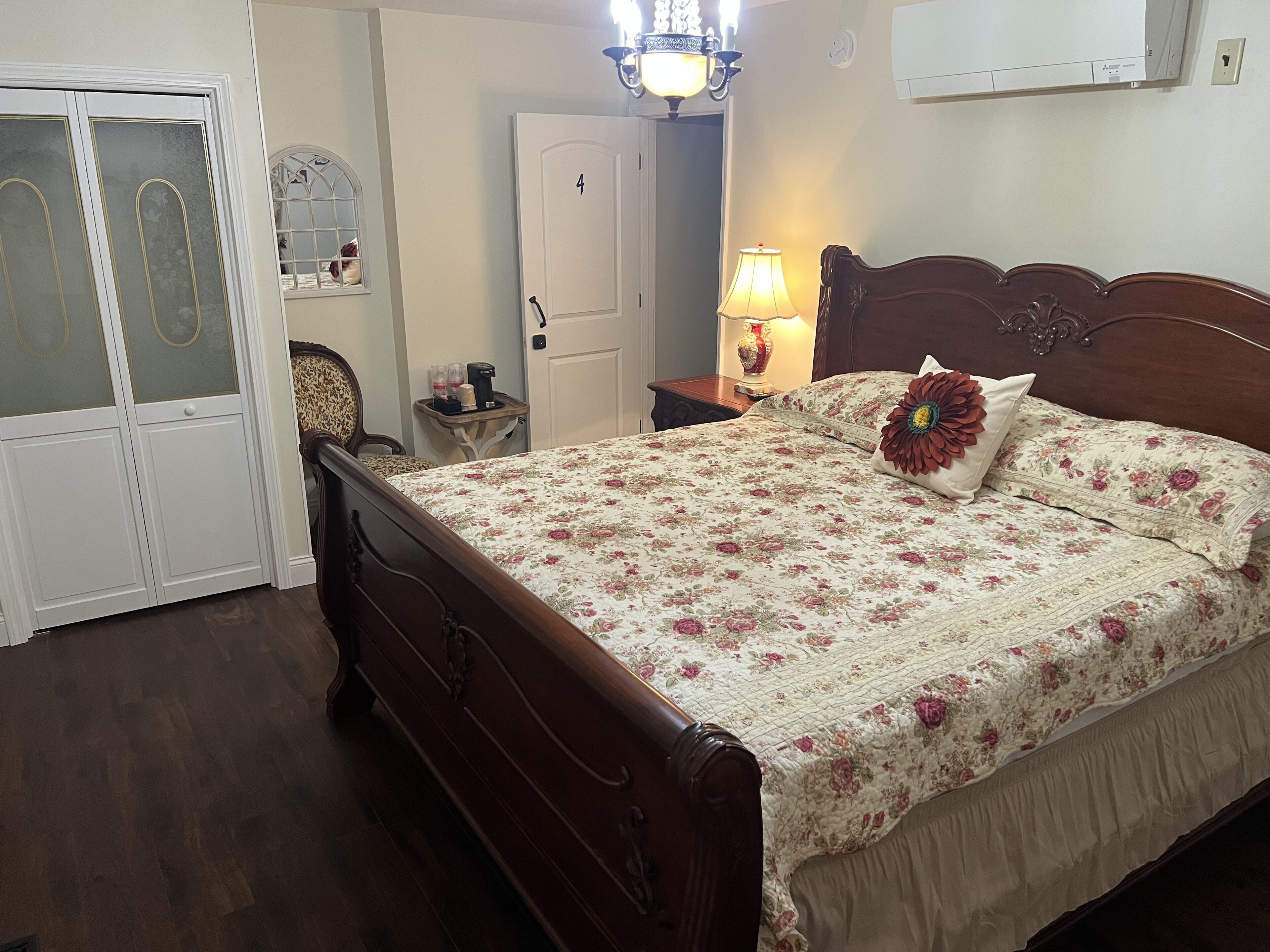 Un dormitorio con cama de madera, ropa de cama floral, mesita de noche con lámpara, armario y una puerta con número.