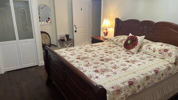 Un dormitorio con cama de madera, ropa de cama floral, mesita de noche con lámpara, armario y una puerta con número.