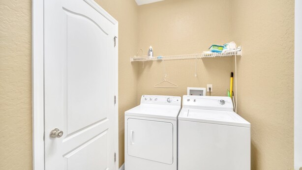 Une buanderie équipée de deux lave-linge et deux sèche-linge à chargement frontal, tous deux blancs, d’une porte blanche et d’une étagère au-dessus des sèche-linge où sont rangés les produits de nettoyage.