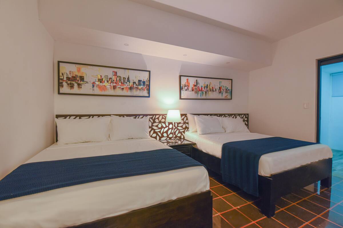 Ein Hotelzimmer mit zwei Betten, einem Nachttisch mit Lampe sowie zwei Stadtansichten an der Wand.